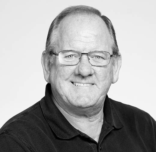 A man with glasses smiles while wearing a dark collared shirt, set against a plain background.