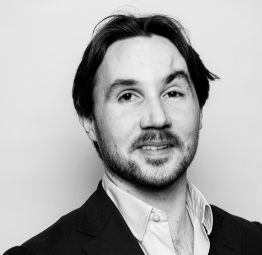 A person with medium-length dark hair and a beard wearing a suit jacket and shirt, smiling slightly, in a black and white portrait.