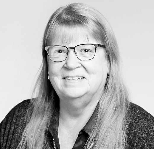 A person with long hair and glasses is smiling in this black and white portrait.
