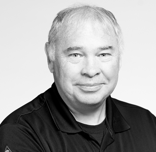 Black and white portrait of an older person with short hair, wearing a dark collared shirt, smiling slightly, and facing the camera.