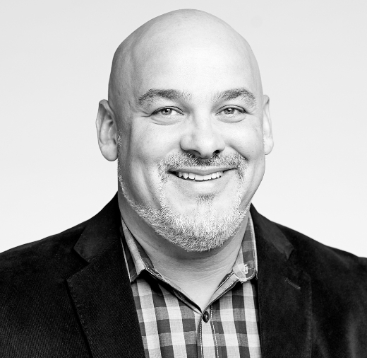 A bald man with a beard smiles while wearing a dark jacket and checkered shirt in a black and white portrait.
