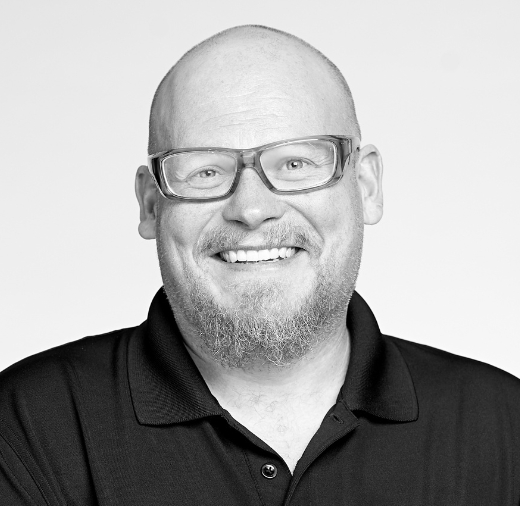 Portrait of a person with glasses, bald head, and a beard, smiling while wearing a black collared shirt against a plain background.