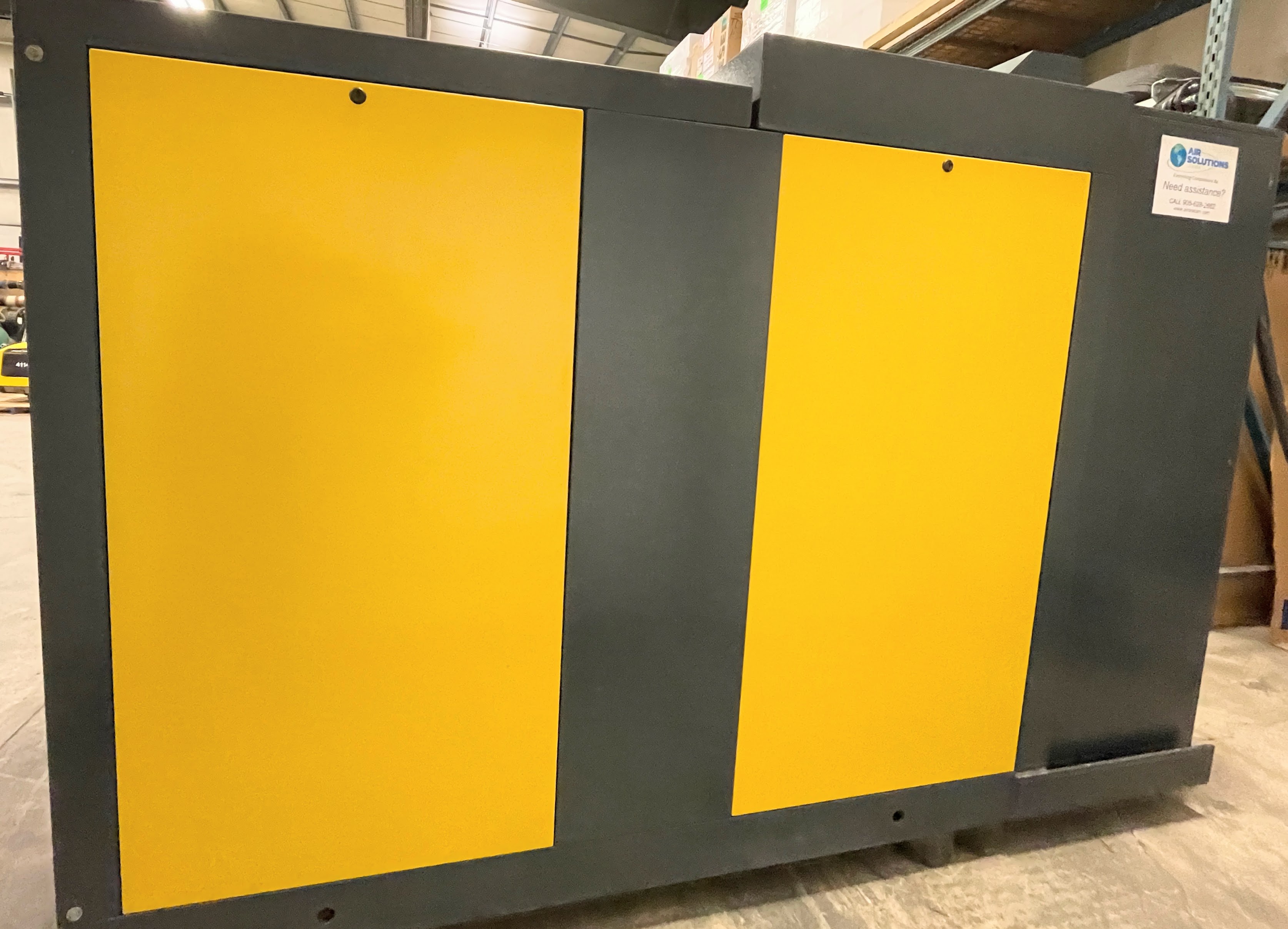 Large industrial machine with yellow and gray panels, placed in a warehouse setting with visible shelves and overhead lighting.