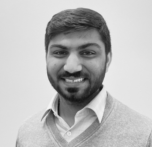 Black and white portrait of a person with a beard, wearing a collared shirt and sweater, smiling at the camera against a plain background.