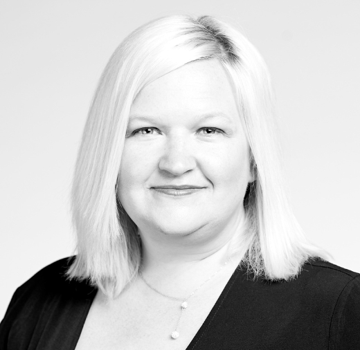 Black and white portrait of a person with medium-length light hair, wearing a dark top and necklace, smiling with a neutral background.