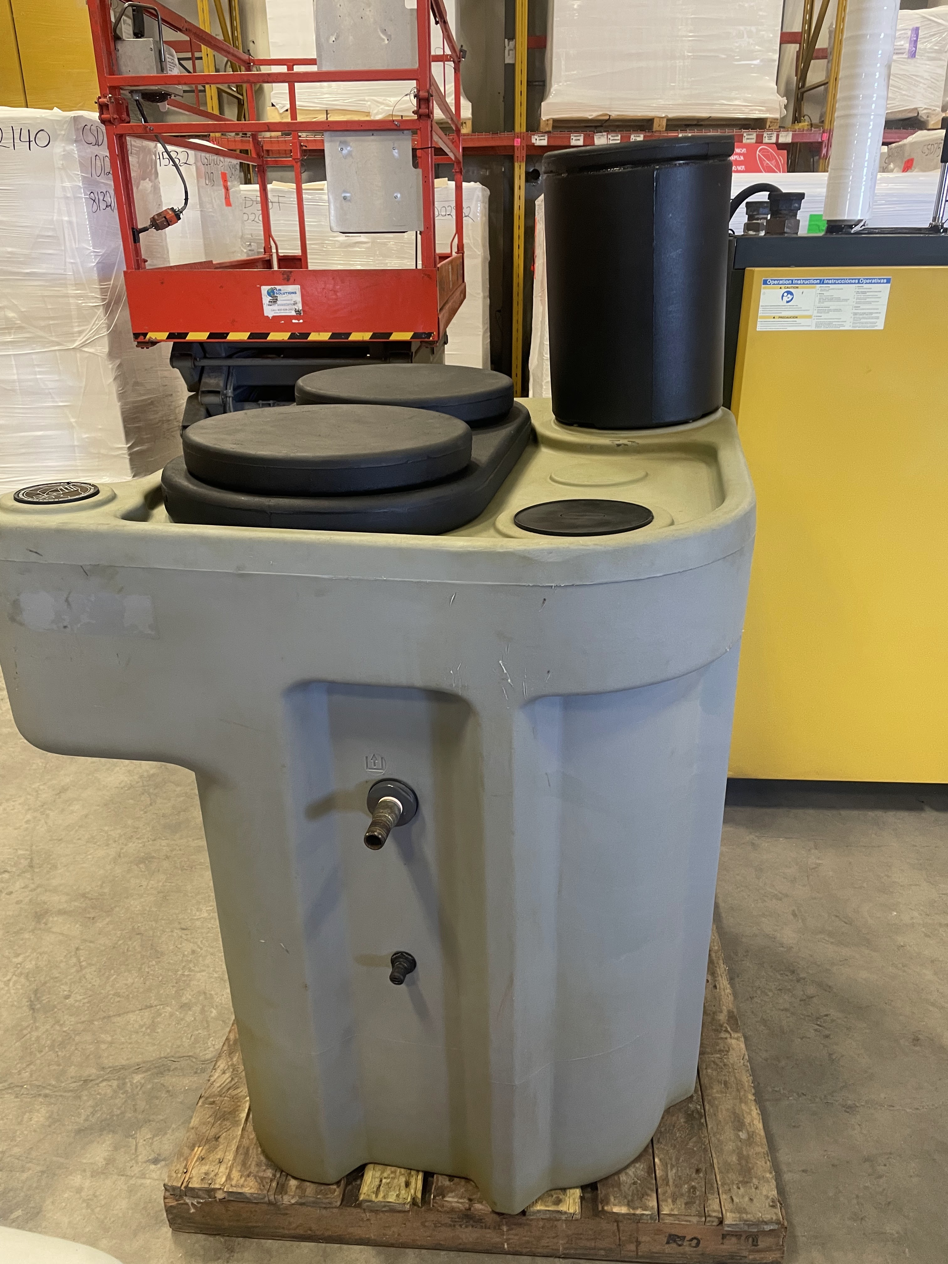 Large gray industrial tank with multiple black lids and a spout, placed on a wooden pallet in a warehouse setting.