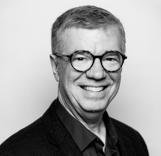 A grayscale photo of a smiling person wearing glasses and a dark blazer.