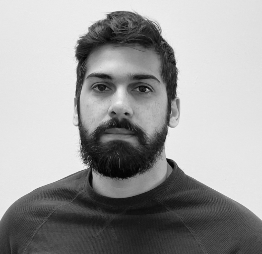 A person with a beard and mustache wearing a dark sweater against a plain background. The image is in black and white.