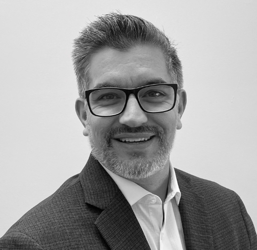 Man with glasses and a beard smiling, wearing a suit and white shirt, in a black and white photo.