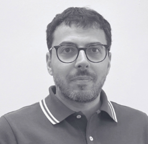 A man with glasses and a beard wearing a collared shirt looks at the camera against a plain background.