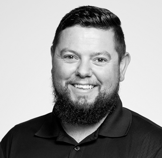 Black and white portrait of a smiling man with a beard, wearing a dark collared shirt, against a light background.