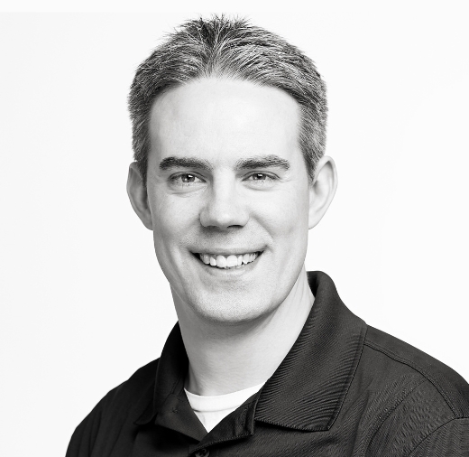 Black and white photo of a smiling person with short hair wearing a collared shirt, looking at the camera against a plain background.