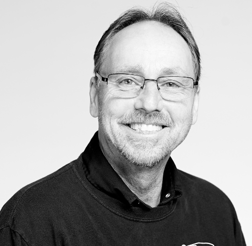 A man with glasses and a beard smiles at the camera in a black and white portrait.