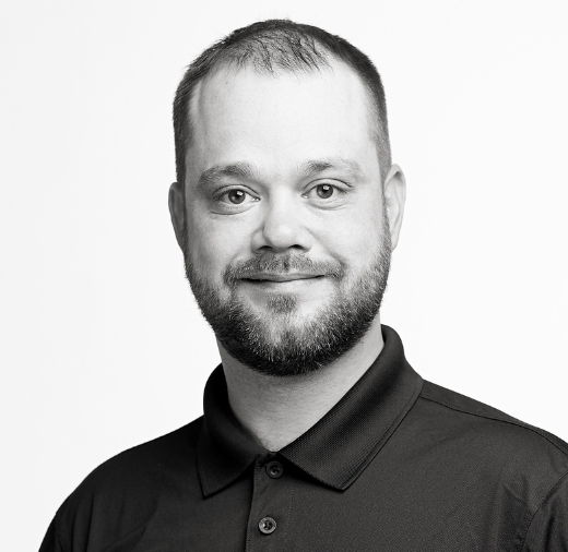 Bearded man in a black shirt smiling, looking directly at the camera, against a plain white background.
