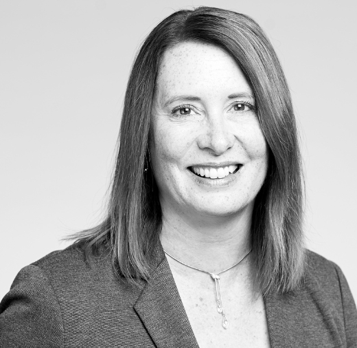 A person with shoulder-length hair smiles in a grayscale portrait, wearing a blazer and a necklace.