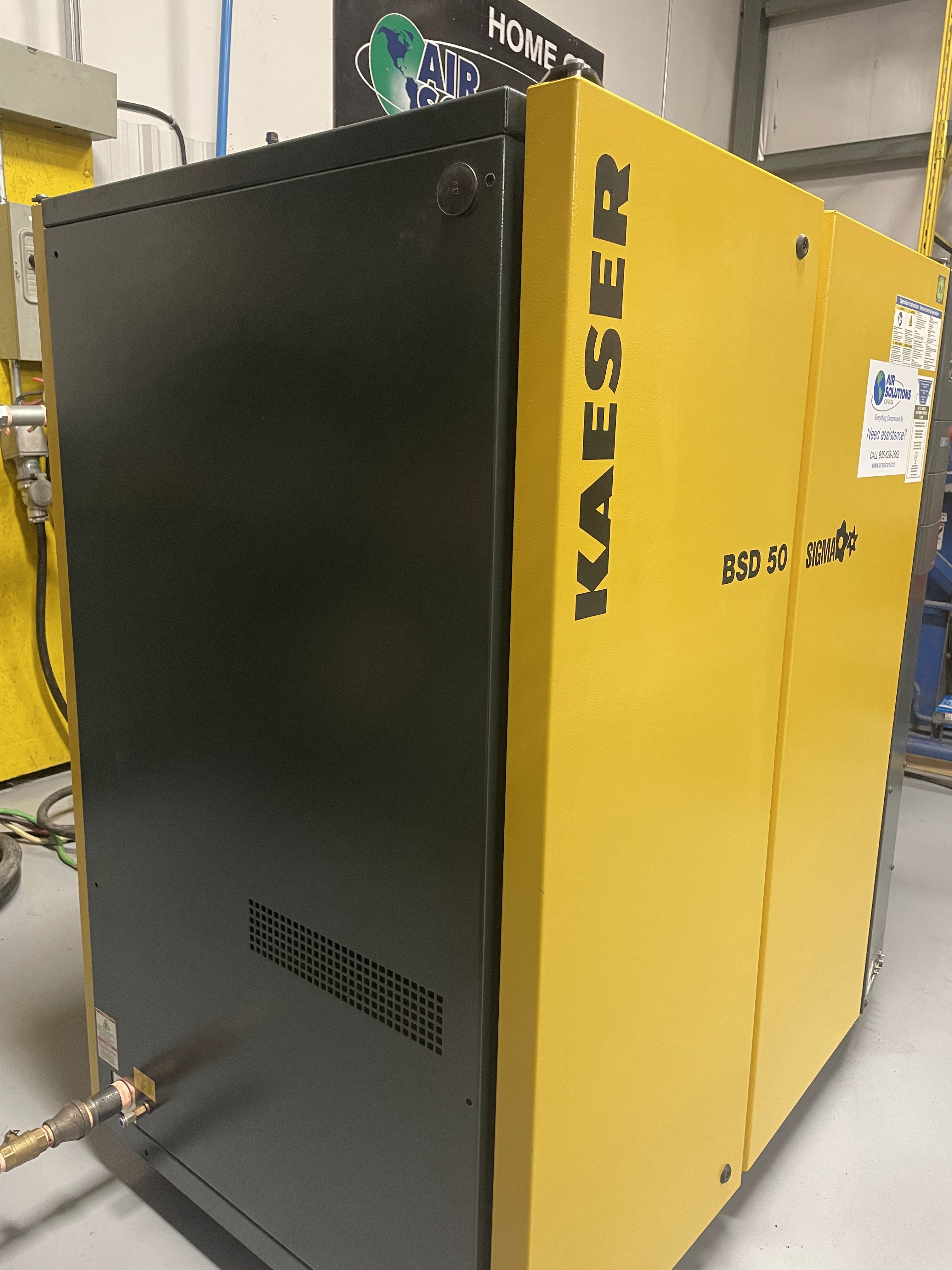 Yellow and black KAESER BSD 50 Sigma air compressor unit is positioned indoors on a gray floor, with visible pipes and control labels.