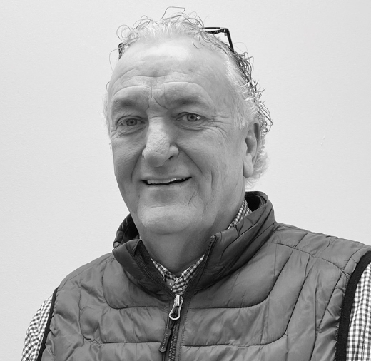 A smiling man with curly hair, wearing a checkered shirt and a quilted vest, stands against a plain background.
