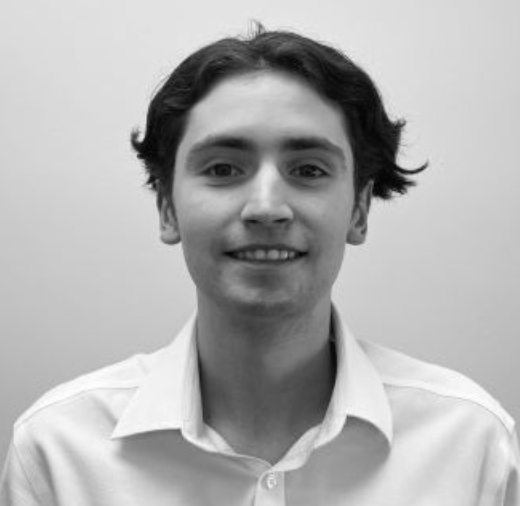 A person with short, dark hair smiles at the camera, wearing a light-colored collared shirt. The background is plain and the image is in black and white.