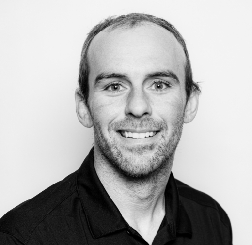 A man with short hair and a beard smiles at the camera in a black and white portrait. He is wearing a dark collared shirt.