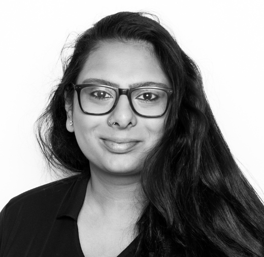 Person with long hair, wearing glasses, and a nose ring, is looking at the camera with a neutral expression. They are wearing a dark collared shirt. The image is in black and white.