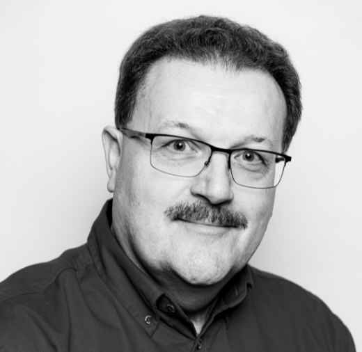 Man with glasses and a mustache wearing a collared shirt, looking at the camera against a plain background. Black and white image.