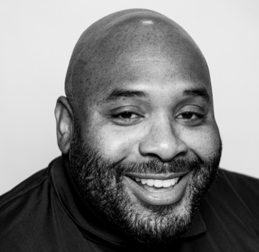 Smiling man in a black shirt, with a shaved head and beard, in a black and white photo.